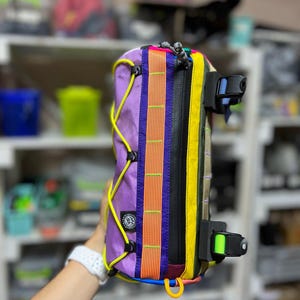 May include: A colorful bicycle frame bag with a purple body, orange and yellow accents, and black zippers. The bag features a variety of straps and buckles in blue, green, and yellow. A yellow cord runs along the side.