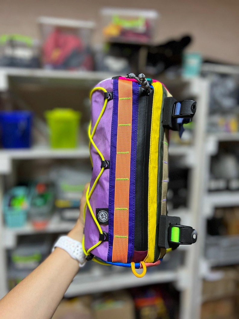 May include: A colorful bicycle frame bag with a purple body, orange and yellow accents, and black zippers. The bag features a variety of straps and buckles in blue, green, and yellow. A yellow cord runs along the side.