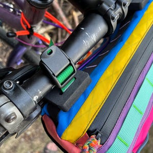 May include: A multi-colored bicycle handlebar bag attached to the handlebars. The bag features a vibrant array of colors including blue, yellow, pink, purple, and green. The bag is secured with a black buckle and strap. The bicycle's handlebars are black.