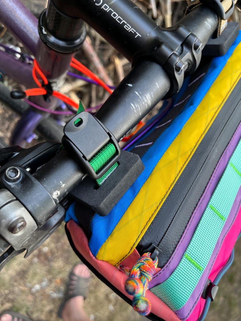 May include: A multi-colored bicycle handlebar bag attached to the handlebars. The bag features a vibrant array of colors including blue, yellow, pink, purple, and green. The bag is secured with a black buckle and strap. The bicycle's handlebars are black.