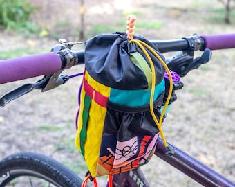 May include: A colorful bicycle handlebar bag with a drawstring closure. The bag is black with panels of yellow, teal, red, and purple. The handlebars have purple grips. The bag is attached to a purple bicycle frame.