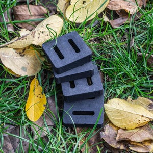 May include: A stack of four dark gray, rectangular foam blocks with two rectangular cutouts each. The blocks are on a bed of green grass, brown leaves, and yellow leaves.