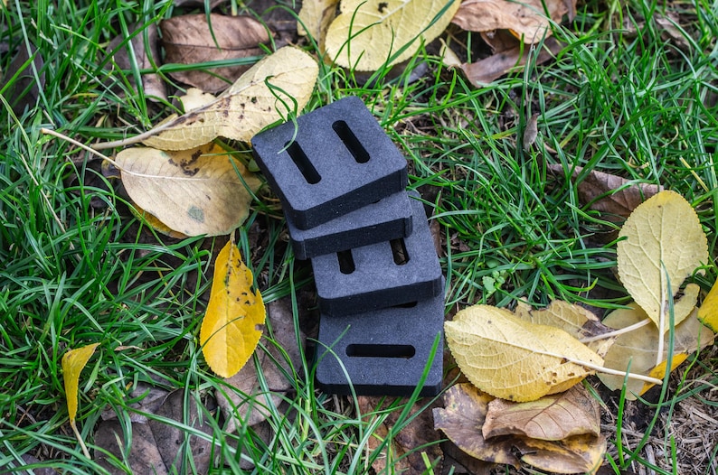 May include: A stack of four dark gray, rectangular foam blocks with two rectangular cutouts each. The blocks are on a bed of green grass, brown leaves, and yellow leaves.