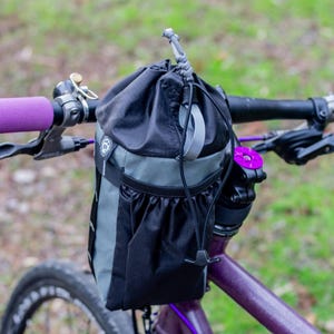May include: A black and grey bicycle handlebar bag with a drawstring closure is mounted on a purple bicycle. The handlebars have purple grips. The bike has a purple frame and a black tyre.