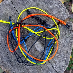 Op de afbeelding: Een verzameling kleurrijke elastische koorden in geel, oranje, blauw en zwart, met zwarte plastic uiteinden. De koorden zijn in een wirwar gerangschikt op een verweerde, grijze boomstronk.