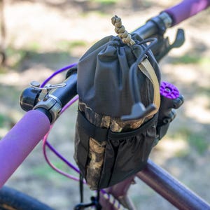 Puede incluir: Una bolsa negra para manillar de bicicleta con un patrón de camuflaje en la sección inferior está sujeta a una bicicleta morada. El manillar tiene puños morados y una campana plateada. La bolsa tiene un cierre de cordón.