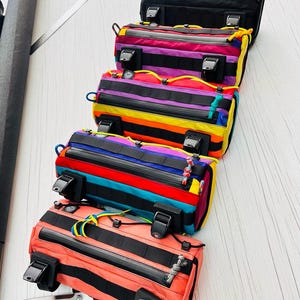 May include: A stack of colorful bicycle handlebar bags. The bags feature a variety of colors, including black, pink, orange, yellow, blue, and purple. Each bag has black straps, zippers, and buckles. The bags are designed for attaching to a bicycle handlebar.