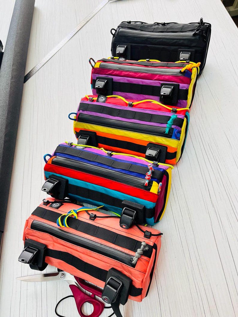May include: A stack of colorful bicycle handlebar bags. The bags feature a variety of colors, including black, pink, orange, yellow, blue, and purple. Each bag has black straps, zippers, and buckles. The bags are designed for attaching to a bicycle handlebar.