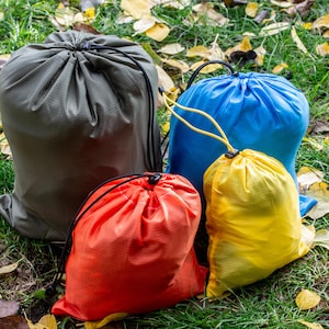 May include: Four drawstring stuff sacks in olive green, red, blue, and yellow are arranged on grass and fallen leaves. The sacks are made of a lightweight material. Each sack has a drawstring closure. The scene is outdoors.