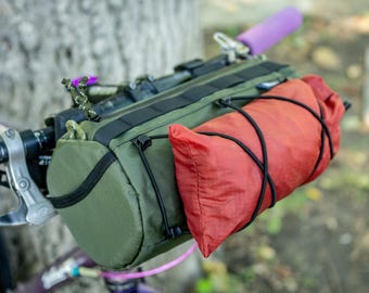 Pode incluir: Um conjunto de guiador de bicicleta com uma bolsa de guiador verde e uma bolsa cilíndrica vermelha, fixadas com cordas elásticas pretas. O guiador tem punhos roxos.