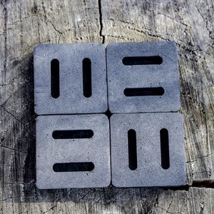 May include: Four square, gray coasters with a textured surface. Each coaster has two parallel, vertical slots or two horizontal slots. The coasters are arranged in a square on a weathered wooden surface.