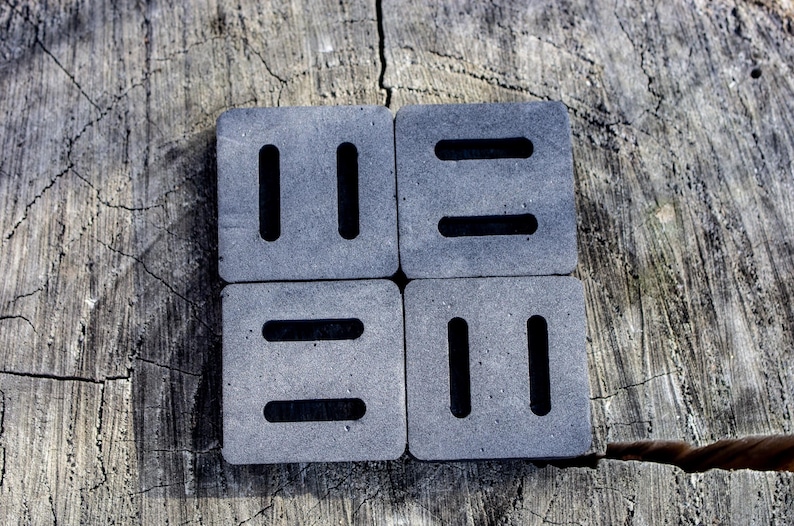 May include: Four square, gray coasters with a textured surface. Each coaster has two parallel, vertical slots or two horizontal slots. The coasters are arranged in a square on a weathered wooden surface.