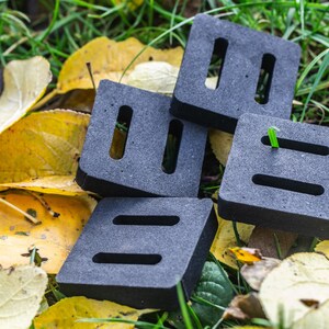 May include: Four gray, square objects with rectangular and oval cutouts are stacked on a bed of green grass and fallen yellow leaves. The objects have a black border. A single blade of green grass is visible.
