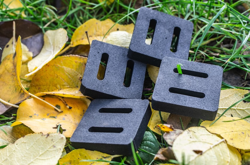 May include: Four gray, square objects with rectangular and oval cutouts are stacked on a bed of green grass and fallen yellow leaves. The objects have a black border. A single blade of green grass is visible.