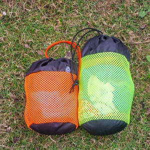 May include: Two drawstring bags on grass. One is orange with a black base and a matching orange handle. The other is green with a black base and a black drawstring. Both bags have a mesh exterior.