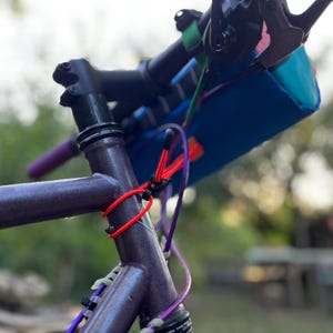 May include: Close-up of a bicycle frame with a purple finish. The handlebars are black with a teal and blue bag attached. Red and purple cords are secured to the frame. The background is blurred with green foliage.