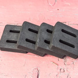 May include: Four dark gray, square coasters with two rectangular cutouts each are stacked on a pink, weathered wooden surface. The coasters appear to be made of a dense, absorbent material.