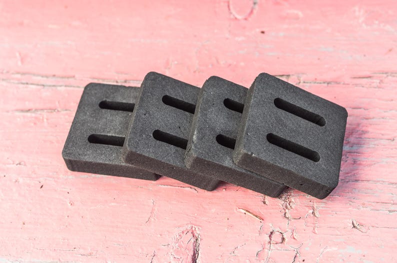 May include: Four dark gray, square coasters with two rectangular cutouts each are stacked on a pink, weathered wooden surface. The coasters appear to be made of a dense, absorbent material.