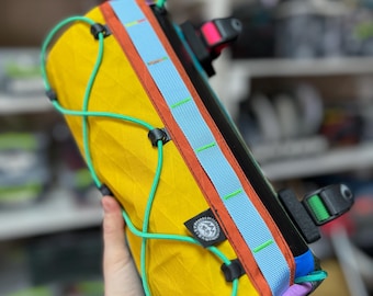 Pode incluir: Uma bolsa cilíndrica para guiador de bicicleta em tecido amarelo-mostarda com padrão de losangos, uma faixa central azul-clara com costuras verdes e um sistema de cordão elástico verde vivo. Inclui um logótipo circular preto e correas pretas.