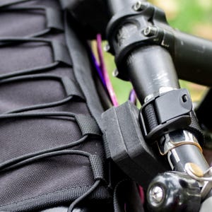 May include: Close-up of a black bicycle handlebar with a mounted black bag and a black strap. The handlebar has a purple accent and a silver bell. The bag has a series of black elastic cords.