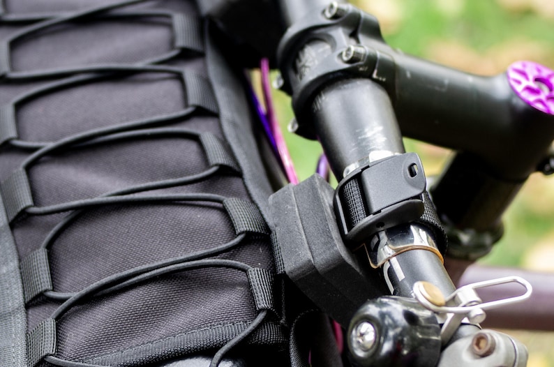 May include: Close-up of a black bicycle handlebar with a mounted black bag and a black strap. The handlebar has a purple accent and a silver bell. The bag has a series of black elastic cords.