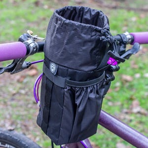 Puede incluir: Una bolsa negra para manillar de bicicleta con cierre de cordón se monta en una bicicleta morada. La bolsa tiene un bolsillo pequeño y se asegura con correas. El manillar tiene puños y manetas de freno morados.