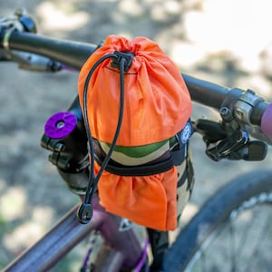 May include: An orange drawstring bag attached to a bicycle handlebar. The bag is mounted on a camouflage-patterned base. The bicycle has purple handlebar grips and a purple stem cap.