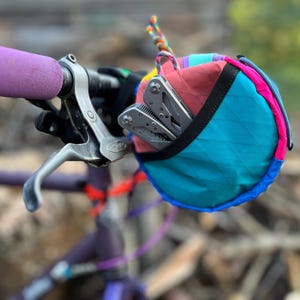 May include: A colorful bicycle handlebar bag with a multi-tool inside. The bag is a mix of teal, pink, red, and blue. The handlebar grip is purple. The multi-tool is silver. The bag is attached to a bicycle handlebar.