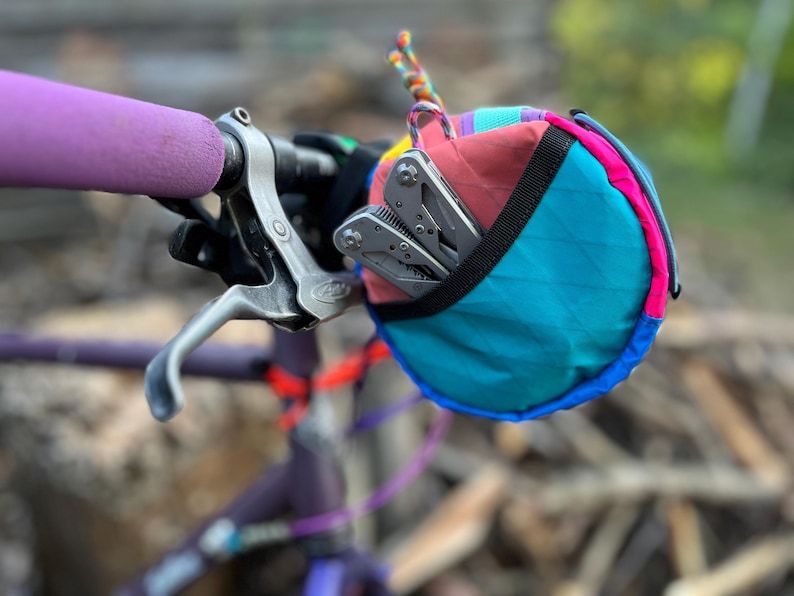 May include: A colorful bicycle handlebar bag with a multi-tool inside. The bag is a mix of teal, pink, red, and blue. The handlebar grip is purple. The multi-tool is silver. The bag is attached to a bicycle handlebar.