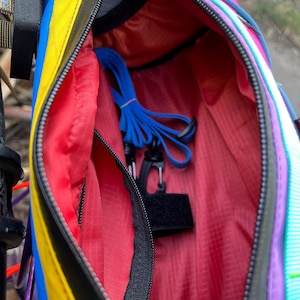 May include: A colorful, open fanny pack with a black zipper and a red interior. The exterior features stripes of yellow, blue, and pink. Inside, a blue strap and black clip are visible. The bag is designed for outdoor activities.