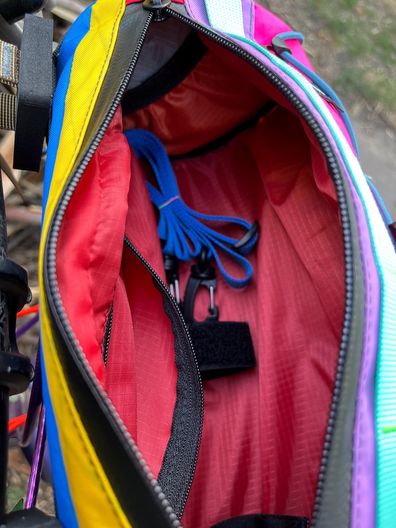 May include: A colorful, open fanny pack with a black zipper and a red interior. The exterior features stripes of yellow, blue, and pink. Inside, a blue strap and black clip are visible. The bag is designed for outdoor activities.