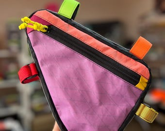Op de afbeelding: Een driehoekige fietstas voor aan het frame met een paars hoofdpaneel voorzien van een subtiel ruitpatroon. De tas heeft een zwarte rits, een feloranje bovenrand en bevestigingsriemen van neongroen, oranje, rood en geel nylon weefsel.