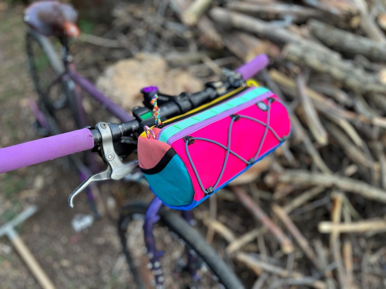 May include: A colorful bicycle handlebar bag in bright pink, teal, and coral, with a black and gray elastic cord design. The bag is mounted on a purple bicycle with purple handlebar grips. The bag has a yellow and teal zipper.