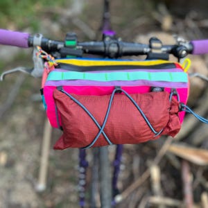 May include: A colorful bicycle handlebar bag with a red pouch and a rainbow-striped top section. The handlebars have purple grips. The bag is attached to the bicycle's handlebars, with a blurred background of natural elements.