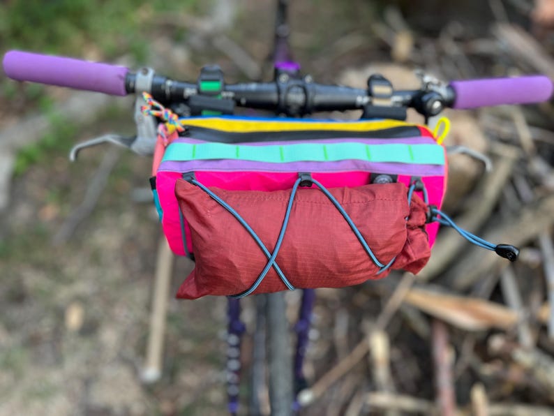 May include: A colorful bicycle handlebar bag with a red pouch and a rainbow-striped top section. The handlebars have purple grips. The bag is attached to the bicycle's handlebars, with a blurred background of natural elements.