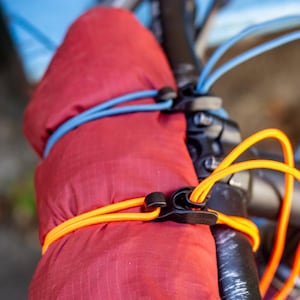 May include: Close-up of a red bicycle handlebar mitt secured with blue and orange bungee cords. The mitt is made of a quilted material. The handlebar and other bike components are black.