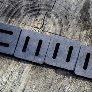 May include: Four dark gray, square foam pads with rectangular cutouts, arranged in a row. The pads are on a weathered, gray wooden surface. The cutouts are evenly spaced and create a pattern.