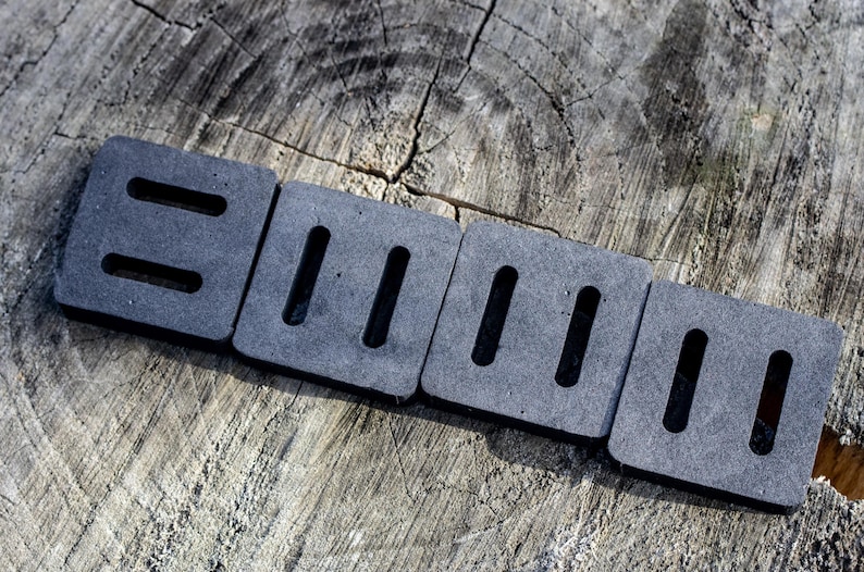 May include: Four dark gray, square foam pads with rectangular cutouts, arranged in a row. The pads are on a weathered, gray wooden surface. The cutouts are evenly spaced and create a pattern.
