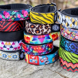 May include: A stack of colorful dog collars with various patterns, including floral, zebra, leopard, geometric, and cartoon designs. Each collar has a black interior and a buckle closure. The collars are stacked on a wooden surface.