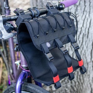 Op de afbeelding: Zwarte fietstas voor het stuur met rode accenten en zwarte riemen. De tas is gemonteerd op een paarse fiets met paarse handvatten. De tas heeft een reeks elastische koorden aan de voorkant.