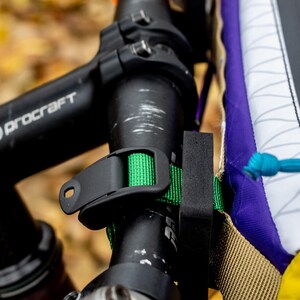 May include: Close-up of a bicycle handlebar with a black Procraft stem and a purple accent. A green strap secures a black accessory to the handlebar. A colorful bag is attached with a blue rope.