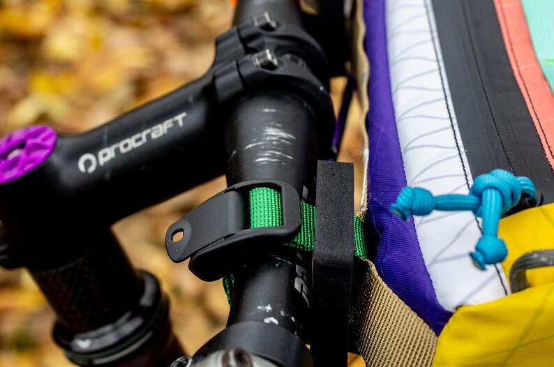 May include: Close-up of a bicycle handlebar with a black Procraft stem and a purple accent. A green strap secures a black accessory to the handlebar. A colorful bag is attached with a blue rope.