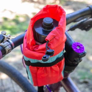 May include: A red and teal bicycle water bottle holder attached to the handlebars. The holder is made of fabric and has a drawstring closure. The handlebars are black with purple grips and a purple accent on the stem.