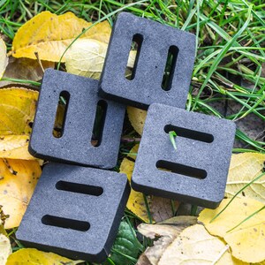 May include: Four gray, square-shaped plant pot feet with rectangular and vertical cutouts are arranged on a bed of yellow leaves and green grass. A small green sprout emerges from one of the feet. The feet are designed to elevate pots.