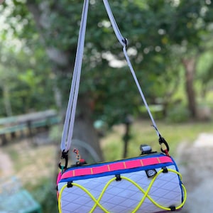 May include: A colorful, cylindrical bag with a white, diamond-patterned body. It features a neon yellow bungee cord design, a pink and blue top, and a white shoulder strap. Black clips and accents complete the design.