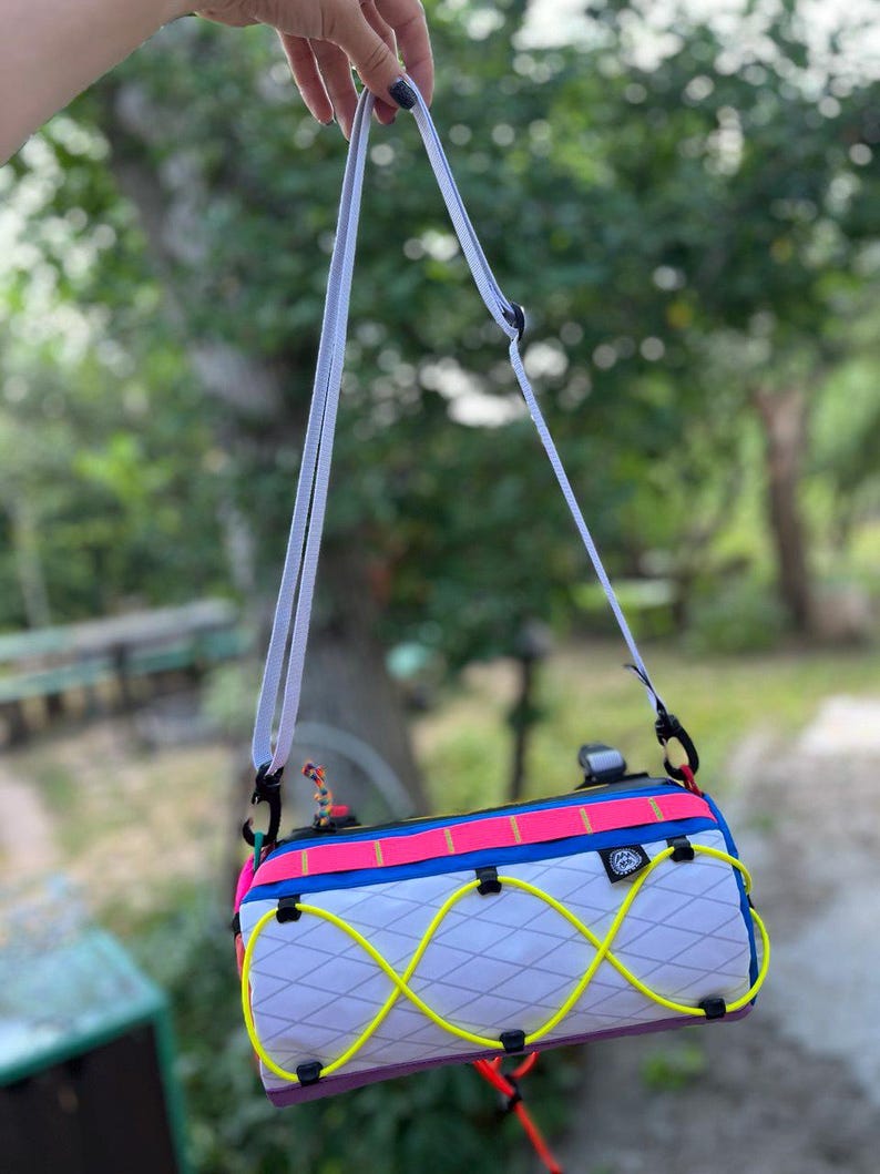 May include: A colorful, cylindrical bag with a white, diamond-patterned body. It features a neon yellow bungee cord design, a pink and blue top, and a white shoulder strap. Black clips and accents complete the design.