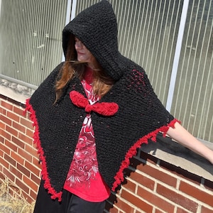 May include: A black hooded cape with red trim and a red bow closure. The cape is made of a thick, textured knit fabric. The edges are trimmed with a red, scalloped design.