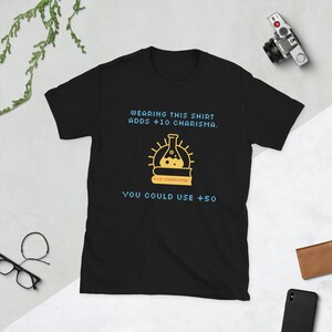 Könnte beinhalten: Schwarzes T-Shirt mit einem Grafikdesign, das einen gelben Becher und den Text "WEARING THIS SHIRT ADDS +10 CHARISMA. YOU COULD USE +50" zeigt. Das T-Shirt besteht aus einem weichen, bequemen Material.