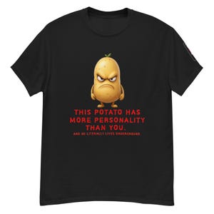 Puede incluir: Camiseta negra con una ilustración de una patata de dibujos animados con expresión enfadada y el texto rojo: "THIS POTATO HAS MORE PERSONALITY THAN YOU. AND HE LITERALLY LIVES UNDERGROUND." La camiseta es de material suave.