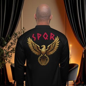 Pode incluir: Casaco bomber preto com acabamento vermelho e um desenho de águia dourada nas costas. O casaco apresenta as letras SPQR em vermelho acima da águia. A águia é rodeada por uma coroa de louros.